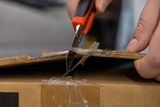 Human hands use a sharp utility knife to carefully cut open a sealed cardboard delivery box, symbolizing e-commerce, logistics, and secure package unpacking