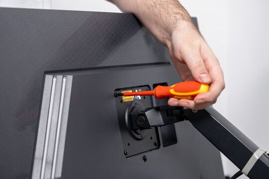 Person's hand uses a precision screwdriver to securely fasten a monitor arm onto a computer display, illustrating practical home office equipment assembly and installation