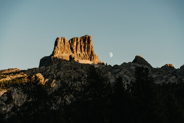 Alpine Berglandschaft im warmen Licht des Sonnenuntergangs, daneben der klar sichtbare Mond am Himmel. Stimmungsvolle Szenerie mit leuchtenden Felsfarben und ruhiger Abendatmosph&auml;re.
