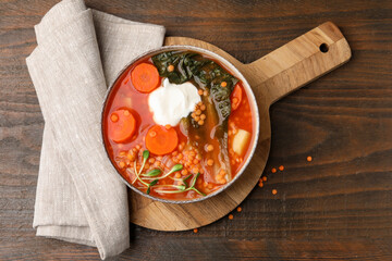 Tasty lentil soup with sour cream and vegetables in bowl on wooden table, top view