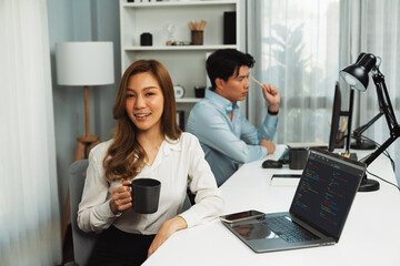 Profile smiling beautiful Asian businesswoman looking camera while holding cup to pose at modern office on working desk. Blurry background man colleague analyzing data market report on pc. Infobahn.