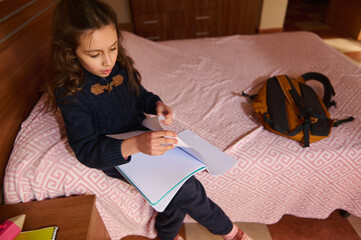 Obraz premium View from above of an elementary age student girl engaged in homework at home, sitting on her bed. A backpack is nearby