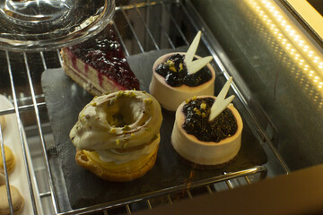 Closeup cheesecake and pistachio pastry display showcasing glossy chocolate sticks, crumbly base, berry tart in background, warm