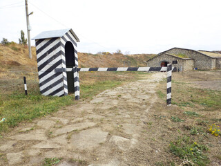 A guard booth painted in black and white stripes. It looks empty and slanted. In front of the booth is a simple wooden barrier, also painted in black and white stripes. It blocks a narrow stone path.