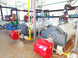 A modern boiler room with two large industrial boilers. The boilers are connected to pipes supplying water and gas. The pipes are equipped with pressure gauges, shut-off valves, pumps, and fittings.