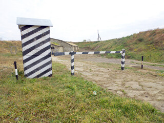 A guard booth painted in black and white stripes. It looks empty and slanted. In front of the booth is a simple wooden barrier, also painted in black and white stripes. It blocks a narrow stone path.