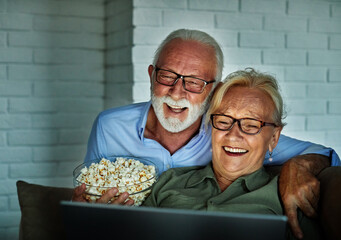 Portrait of an elderly senior happy couple using laptop at night, using modern technology having fun with glowing screen in dark office or at home, watching tv together eating popcorn at home. Shot of