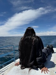 A young woman with black, long hair sits on the deck of a tourist boat while she looks to the beautiful Titicaca-lake in Peru.