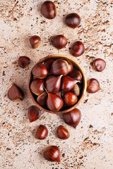 Wooden bowl filled with chestnuts on beige stone kitchen table, autumn seasonal food themes, top view