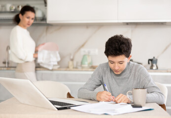 Teen son doing school homework while mom washes dishes in the kitchen