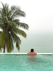 it is raining heavily in the background, a man in the pool is relaxing and watching the rain in the sea, a palm tree by the pool in a luxury villa and the sea