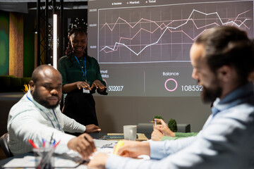 Black executive assistant developing a business strategy and goals, studying market trends and dynamics shown on projection board. Group of workers attending a night time meeting.