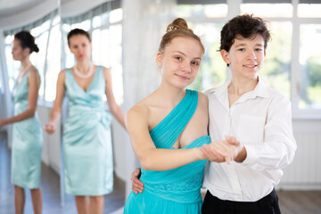 Passionate young ballroom enthusiasts, teenage girl and boy practicing graceful waltz routine in well-lit dance hall under female mentor watchful eye