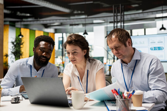 Multiethnic corporate team, including black man and caucasian individuals, collaborating in office, using laptop and documents to assess investment reports and business analytics. Teamwork in action. - Powered by Adobe