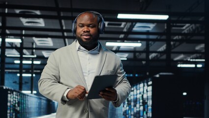 Data center admin wearing headphones using tablet to identify bottlenecks. Server farm IT expert listening music while troubleshooting bugs, implementing optimizations, camera B