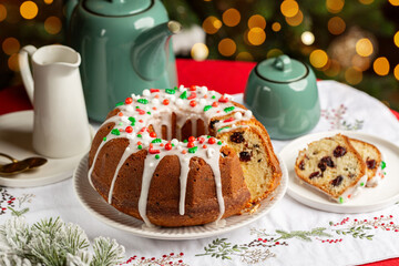 Festive table with homemade pastry, Christmas or New Year bund cake with cranberries, sugar icing and decoration, tea pot and mugs. Christmas tree and lights on background.