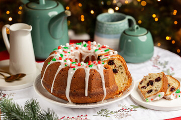 Served Festive table with homemade pastry, Christmas or New Year bund cake with cranberries, sugar icing and decoration, tea pot and mugs. Christmas tree and lights on background.