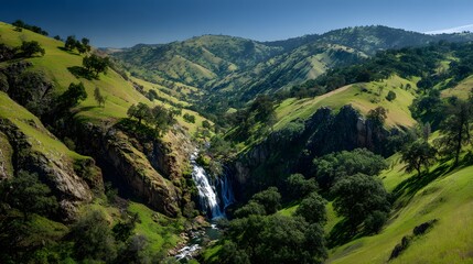 Fototapeta premium Lush green rolling hills cradle a cascading waterfall flowing through a deep ravine under a clear blue sky