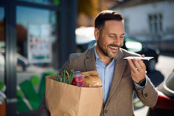 Happy man talking over cell phone's speaker while leaving supermarket with full grocery bag. © Drazen