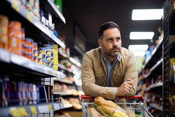 Displeased man leaning on shopping cart while buying at supermarket.
