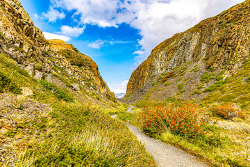 A narrow hiking trail winds through steep rocky canyon walls surrounded by rugged vegetation in Torres del Paine National Park. © Zhiqi