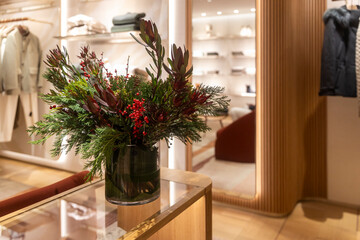 A festive arrangement of green pine foliage, red berries, and deep red flowers sits in a glass vase on a wooden and glass table inside a brightly lit, modern luxury clothing store.