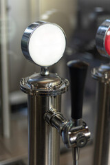 Bright stainless-steel beer tap handle in a craft brewery, with the illuminated round label holder in focus and blurred industrial brewing equipment in the background.