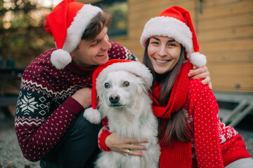 Smiling couple in Santa hats posing with their white dog dressed for Christmas. Festive outdoor holiday portrait with cozy sweaters and cheerful winter atmosphere.