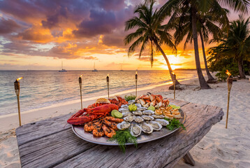 Sunset Seafood Feast - a large platter of lobster, crab, and oysters sits on a wooden table on a tropical beach at sunset.
