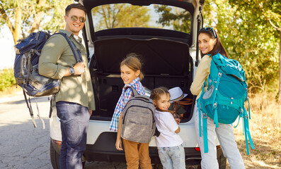 Happy family with backpacks by car trunk preparing for road trip. Parents and children pack luggage for outdoor travel and vacation on a sunny road near countryside. Concept family adventure.