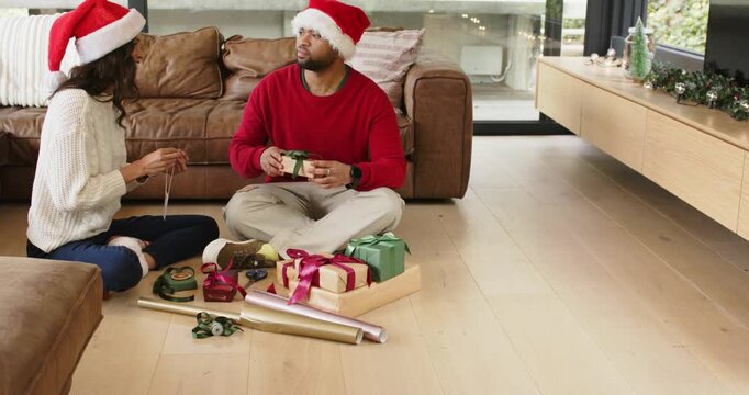 Diverse couple wearing Santa hats threading gift tag before wrapping holiday gifts in living room