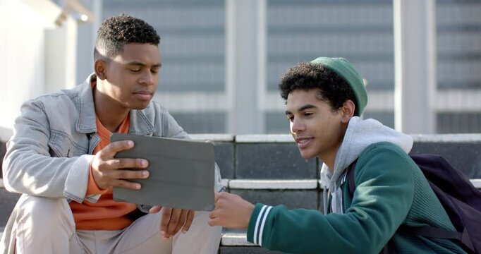 Diverse male friends pointing then rotating tablet on steps for clear view, sharing laughter