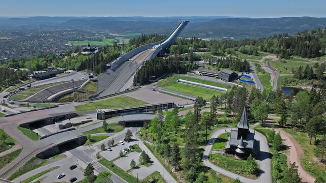 4K Drone View of Holmenkollen Ski Jump, Norway. Tourists visiting the scenic stadium and tower viewpoint in summer daylight, aerial camera glide over curved roads, pine forest, and distant city.