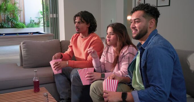 Three friends eating popcorn from striped boxes while reacting surprisingly to movie scene on couch