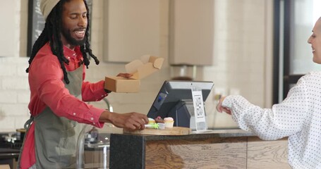African American male barista boxing cupcakes in bakery as customer paying with QR code, copy space