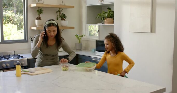 Mother and daughter taking call at kitchen island placing grapes and juice in backpack for school
