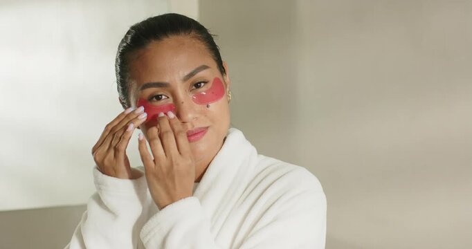 Once red gel under-eye patches applied, Asian woman in robe tapping, swiping at vanity for adhesion