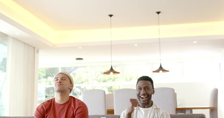 Upon scoring point, diverse male friends are pumping fists, sliding onto grey sofa with controllers