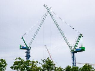 Two tall green construction cranes with angled arms forming an arch shape against a cloudy sky, surrounded by trees in the foreground.