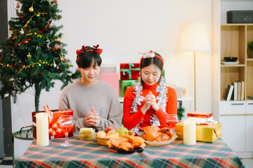 Smiling Asian couple celebrating Christmas together, wearing Santa hats and taking a romantic selfie at a festive dinner table with gifts and candles.