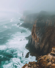 High cliffs above turquoise sea fade into heavy fog. A dramatic coastal landscape with waves crashing against rocks, captured in soft cold tones