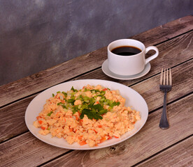 Delicious omelette with tomatoes, decorated with chopped green onions and parsley on a wooden table, a fork and a cup of black coffee nearby.