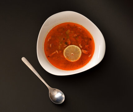 Solyanka, Russian delicious soup in a deep bowl with a slice of lemon on a black background, a tablespoon nearby.