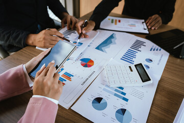A group of professionals reviews charts on paper and a tablet, discussing financial data and calculating figures during a business meeting.