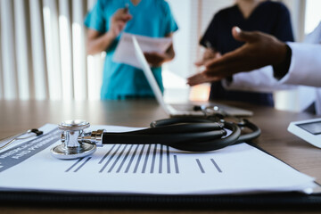 A stethoscope lies on a wooden desk while medical professionals work on a laptop in the background during a healthcare meeting.