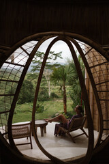 A woman relaxes in a round bamboo archway overlooking lush greenery and tropical scenery at an eco villa