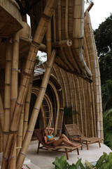 A woman rests on a lounge chair outside a bamboo eco villa overlooking lush tropical greenery and handcrafted wooden architecture