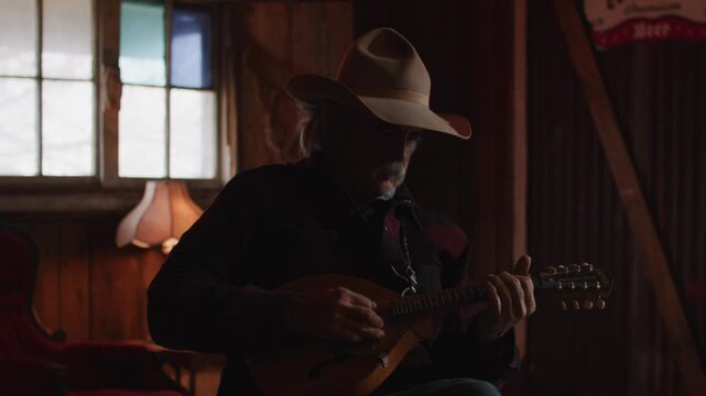 Man With Mustache Wearing Cowboy Hat Playing Mandolin String Instrument Inside of Shadowy Saloon
