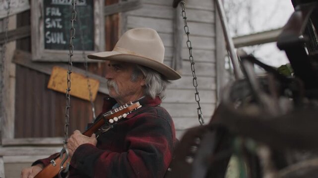 Man Wearing Cowboy Hat and Flannel Shirt Holding Mandolin String Instrument and Sitting on Porch Swing Outside of Old Rustic Saloon