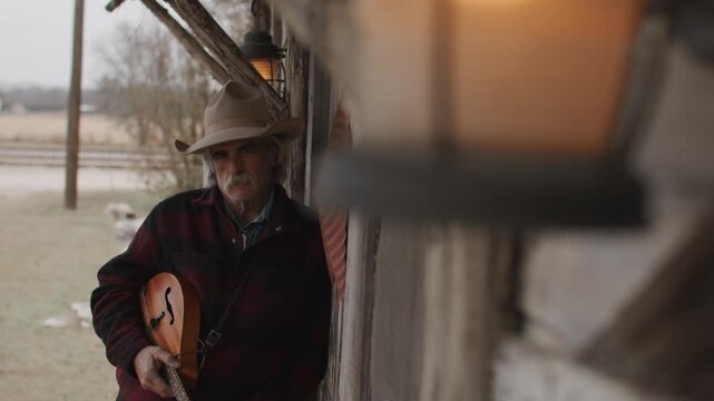 Man Wearing Cowboy Hat and Flannel Shirt Holding Mandolin String Instrument Leaning Against Outside of Old Rustic Saloon